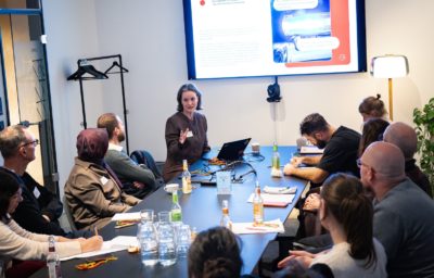 A woman leads a meeting at a long conference table, speaking and gesturing while seated participants listen and take notes. A presentation slide is displayed on a wall-mounted screen behind her. The room is modern and well-lit, with laptops, notebooks, water bottles, and coffee cups spread across the table.
