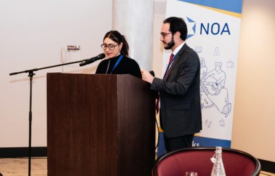 Two people stand at a podium on a stage, one speaking into a microphone while the other looks on. A banner with logos and text hangs behind them, and rows of empty chairs are visible in the foreground, suggesting a formal presentation or conference setting.